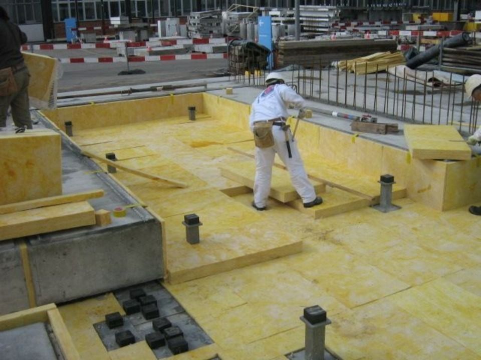 Ein Bauarbeiter in weisser Arbeitskleidung montiert gelbe Dämmplatten auf einem erhöhten Bodengitter auf einer Baustelle.