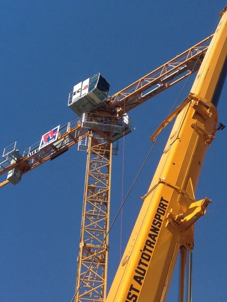 Ein mobiler Kranausleger vor einem hohen gelben Turmdrehkran vor klarem blauen Himmel.