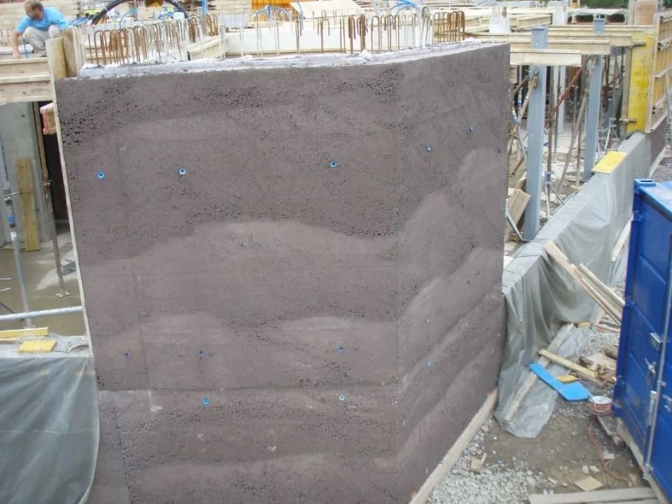 Eine teilweise errichtete Betonwand mit sichtbaren horizontalen Schichten und eingebetteten blauen Befestigungselementen auf einer Baustelle.