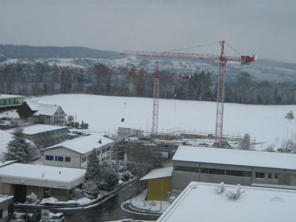 Eine schneebedeckte Baustelle mit zwei roten Kränen, umgeben von Gebäuden und einem Feld unter bedecktem Himmel.