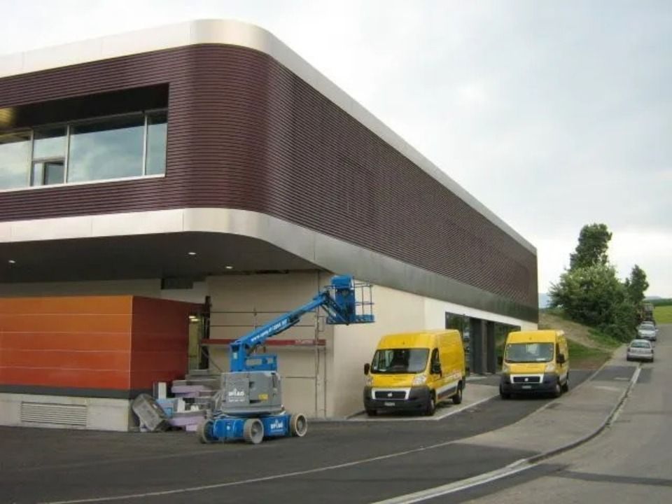 Eine blaue Hubarbeitsbühne stand in der Nähe eines zweistöckigen modernen Gebäudes mit orangefarbenen Verkleidungen, daneben parkten zwei gelbe Lieferwagen.