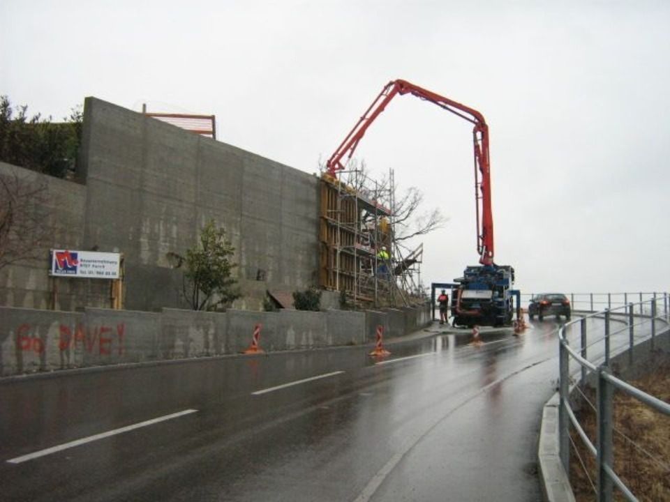 Ein Betonpumpenwagen fährt seinen Ausleger aus, um Zement hinter einer Stützmauer entlang einer nassen Strasse zu verfüllen.