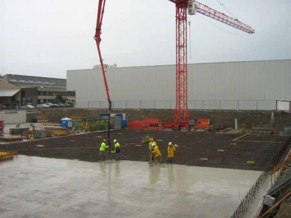 Bauarbeiter in Warnwesten giessen Beton auf einer Baustelle mit einem hohen roten Kran und einem grossen weissen Gebäude.