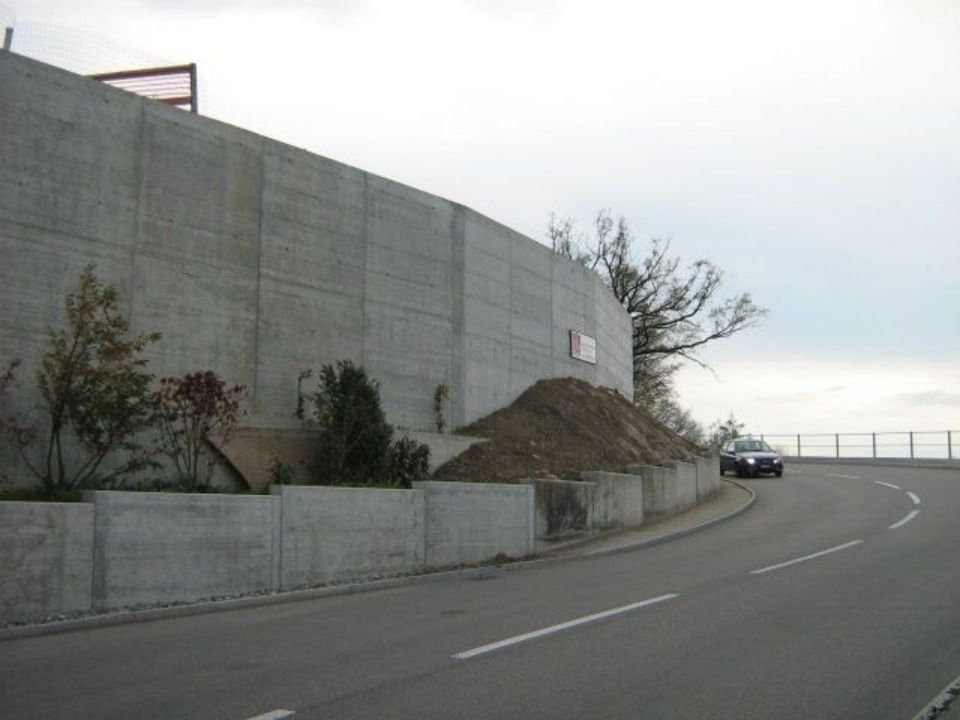 Eine asphaltierte Strasse schlängelt sich an einer hohen Betonstützmauer vorbei, an deren Fuss sich ein kleiner Erdhaufen und einige wenige Büsche befinden.