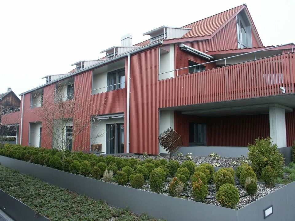 Im Vordergrund ein rotes, mehrstöckiges Wohngebäude mit schrägem Dach, Balkonen und einer akkurat gestutzten Hecke.
