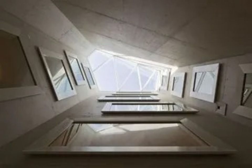 Eine Ansicht aus der Froschperspektive auf einen schmalen, mit gerahmten Spiegeln ausgekleideten Betonkorridor, der zu einem hellen, geometrischen Oberlicht führt.