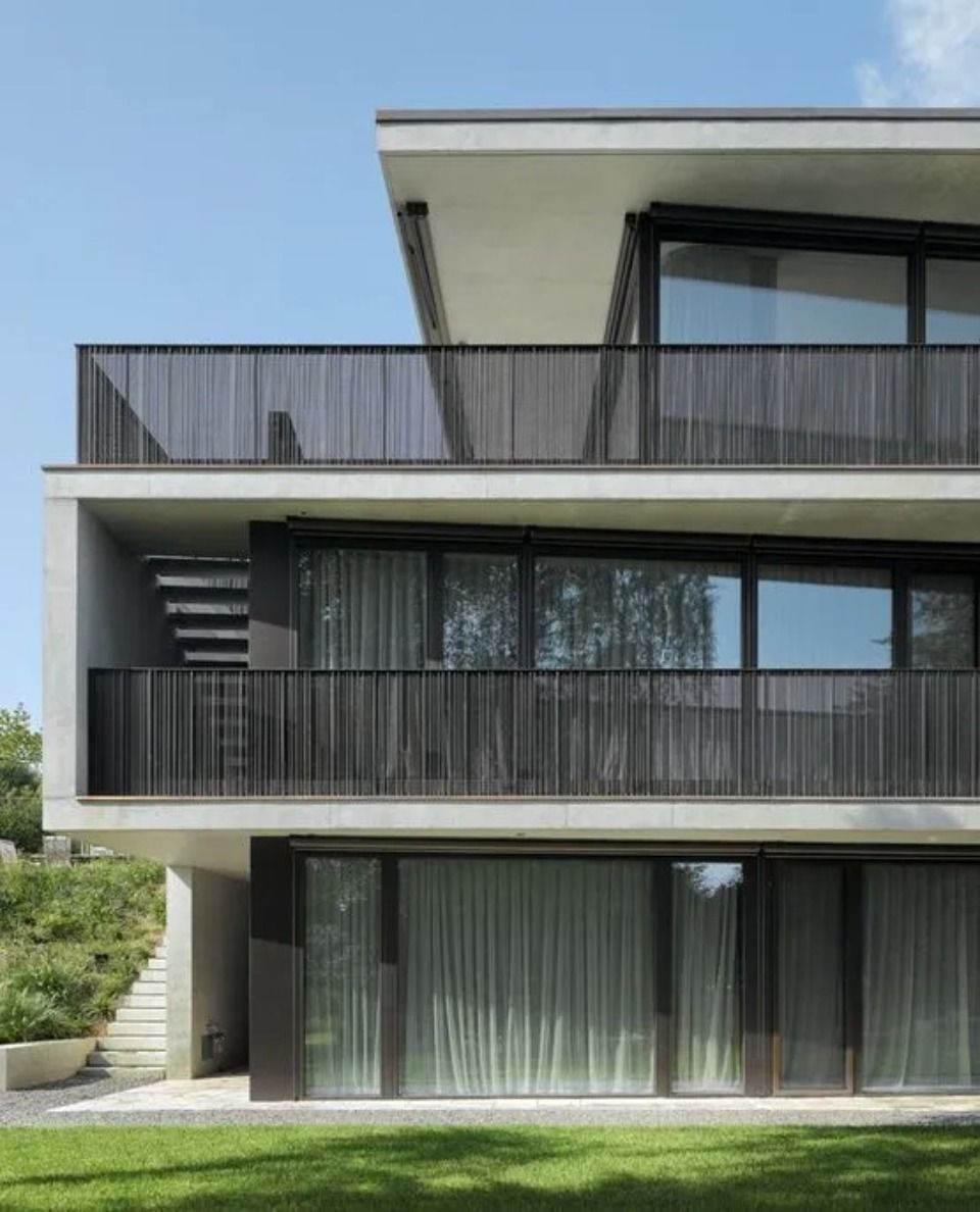 Ein modernes dreistöckiges Gebäude mit weissen Betonterrassen, dunklen Metallgeländern und grossen Glasfenstern auf einer Rasenfläche.