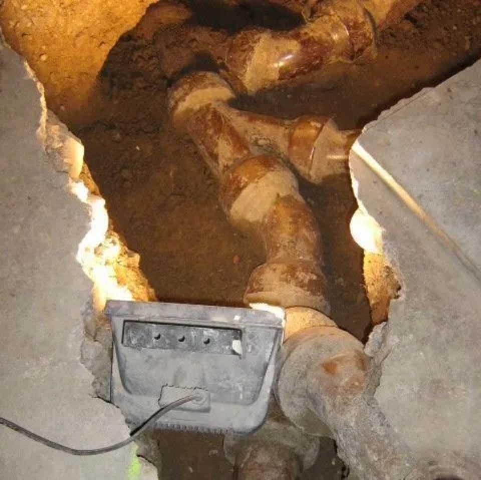 Eine Arbeitsleuchte beleuchtet braune Abflussrohre, die in einem schmalen, durch den Betonboden geschnittenen Graben freiliegen.