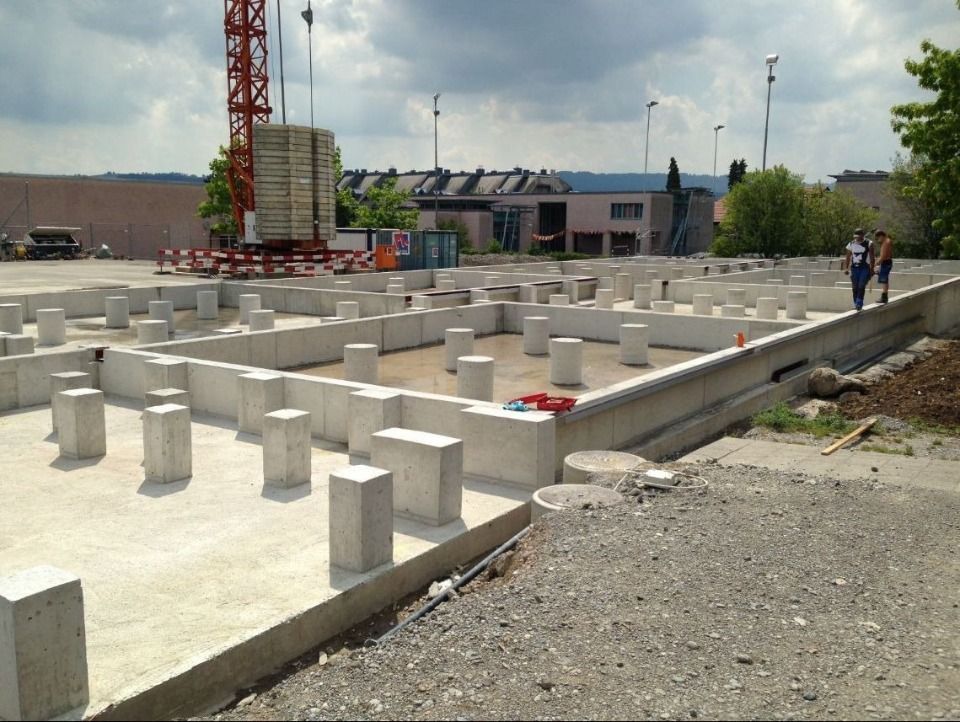 Betonfundamente für ein im Bau befindliches Gebäude, im Hintergrund ein hoher Kran unter bewölktem Himmel.