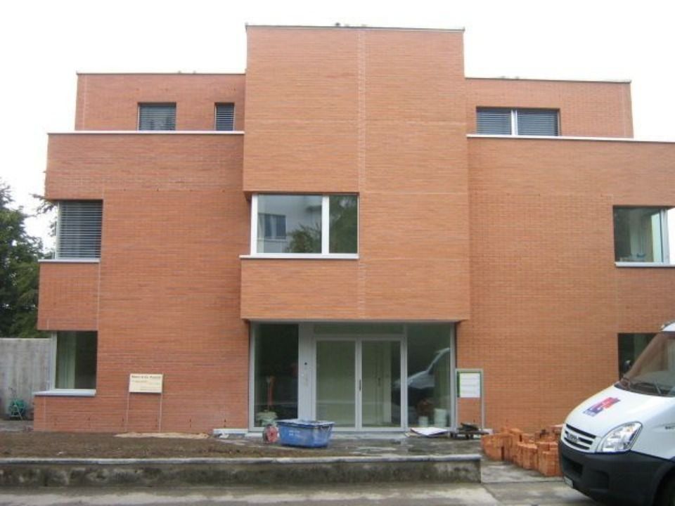 Ein dreistöckiges, modernes Gebäude aus orangefarbenen Ziegeln mit rechteckigen Fenstern und gläsernen Eingangstüren, von der Strasse aus gesehen.