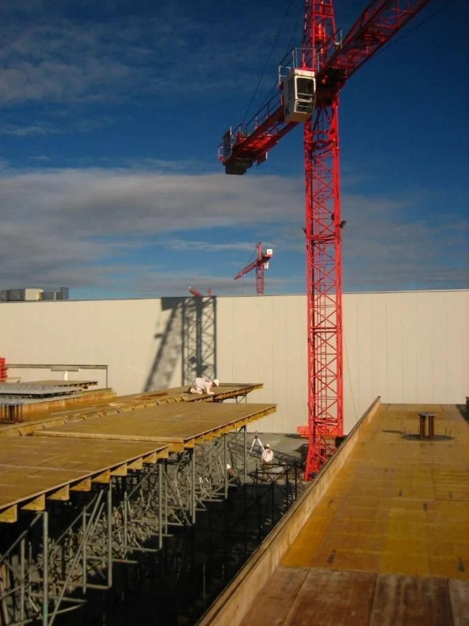 Ein hoher roter Kran ragt über eine Baustelle mit Holzplattformen und Metallgerüsten vor blauem Himmel.