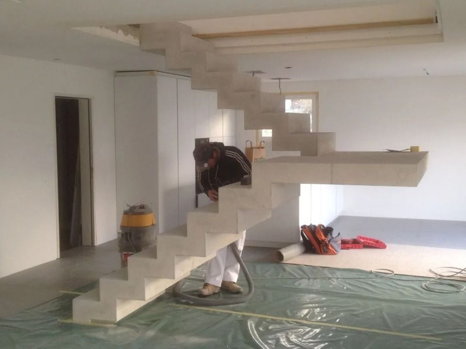 Ein Arbeiter in weissen Hosen reinigt mit einem Staubsauger eine neu installierte, moderne freitragende Betontreppe in einem Innenraum.