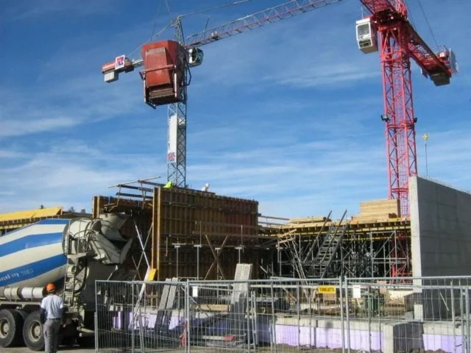 Ein roter Baukran ragt über eine Baustelle mit Holzschalung, Gerüsten und einem geparkten Betonmischer.