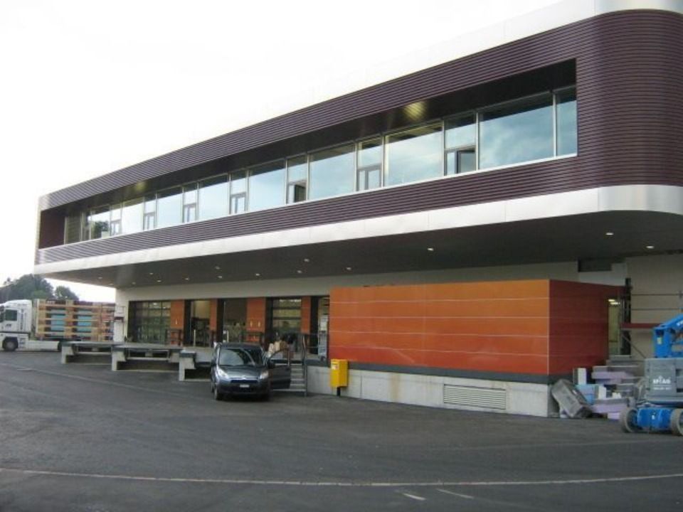 Ein modernes Geschäftsgebäude mit einem orangefarbenen Erdgeschoss und einem auskragenden zweiten Stockwerk mit wellenförmiger brauner Verkleidung.