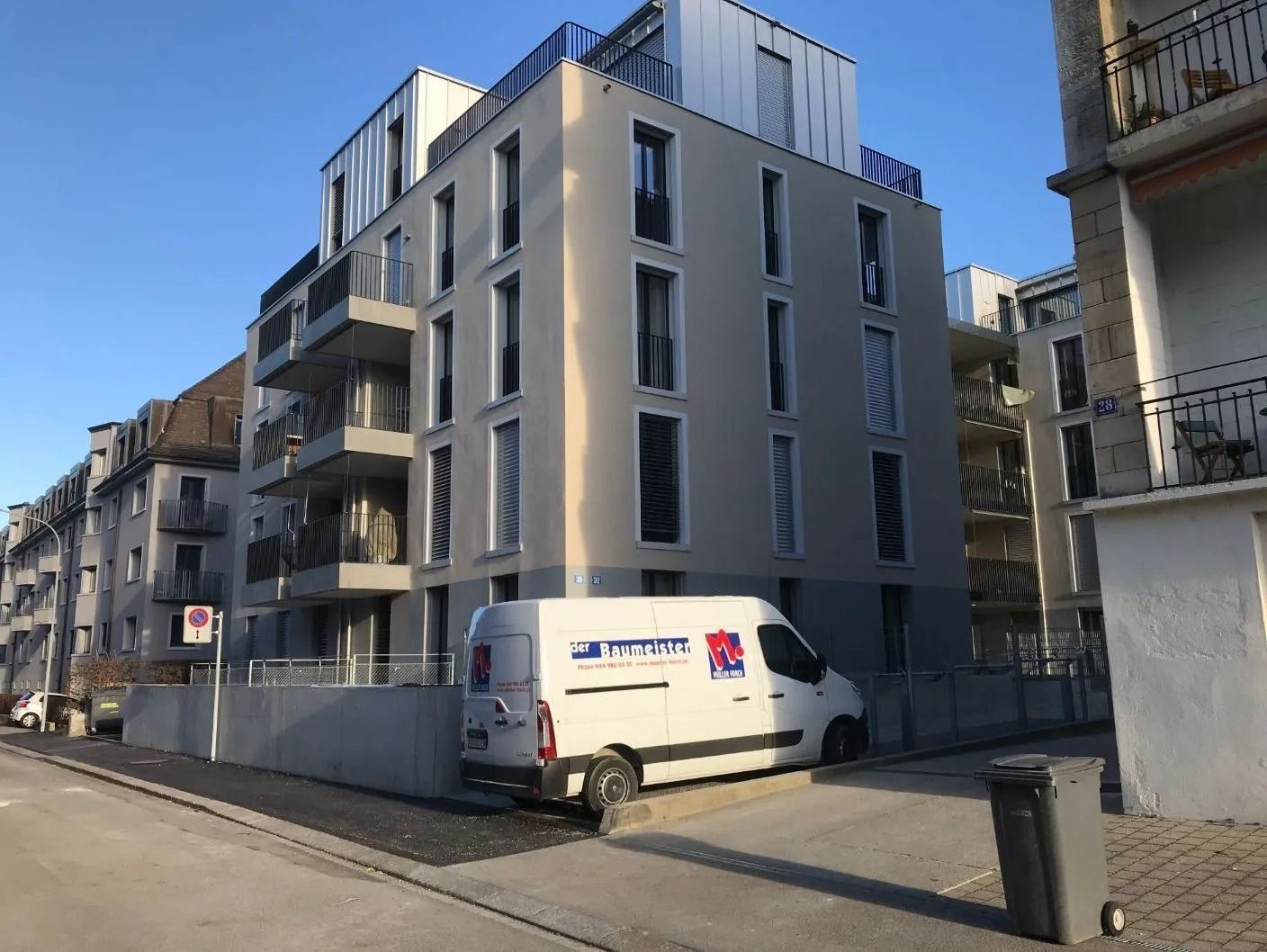 Ein modernes, mehrstöckiges beigefarbenes Wohnhaus mit Balkonen und einem weissen Lieferwagen, der an einem sonnigen Tag davor parkt.