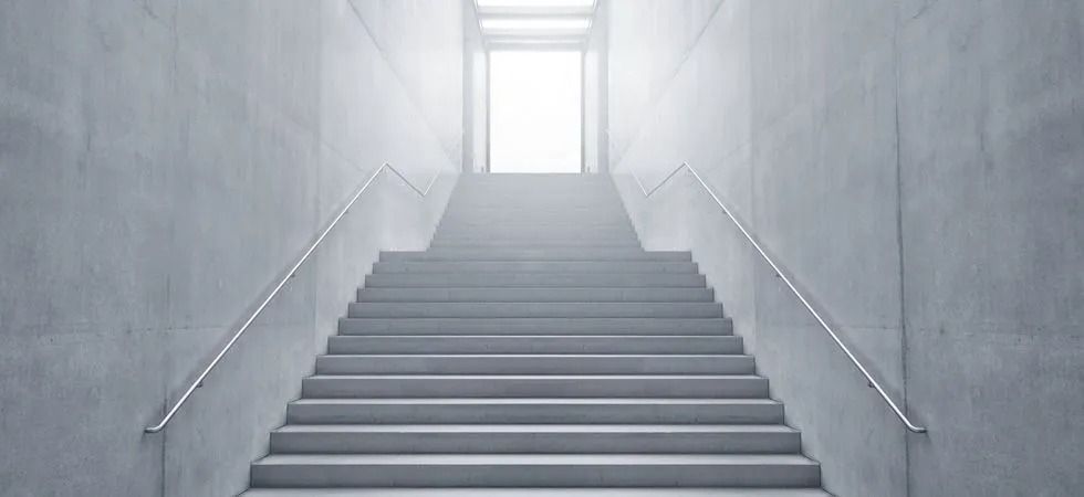 Eine Betontreppe mit silbernen Handläufen führt nach oben zu einem hellen, leuchtenden Licht an der Spitze.