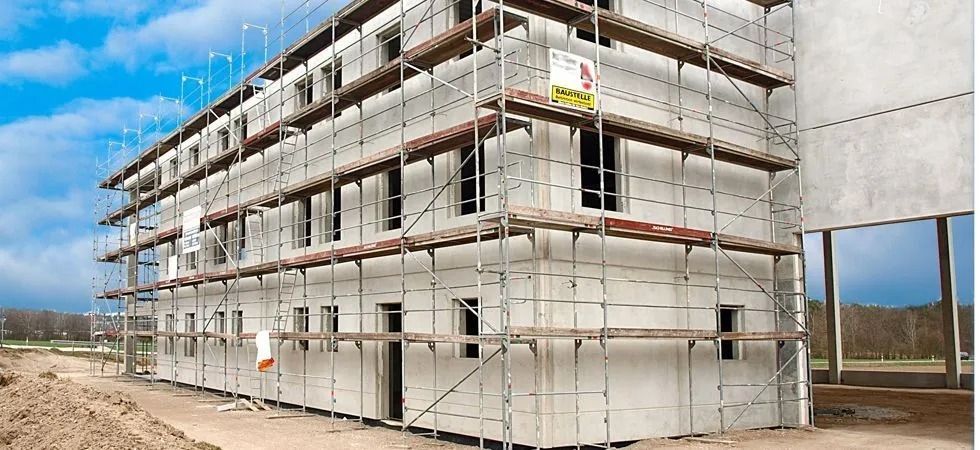 Auf einem unbefestigten Grundstück befindet sich ein mehrstöckiges, im Bau befindliches Gebäude, das mit weissen Paneelen verkleidet und von einem Metallgerüst umgeben ist.