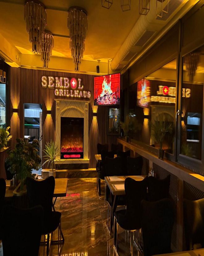 Das Interieur eines eleganten, warm beleuchteten Restaurants ist mit Tischen, Stühlen, Pflanzen und einem leuchtenden Kamin in der Nähe eines Fensters ausgestattet.