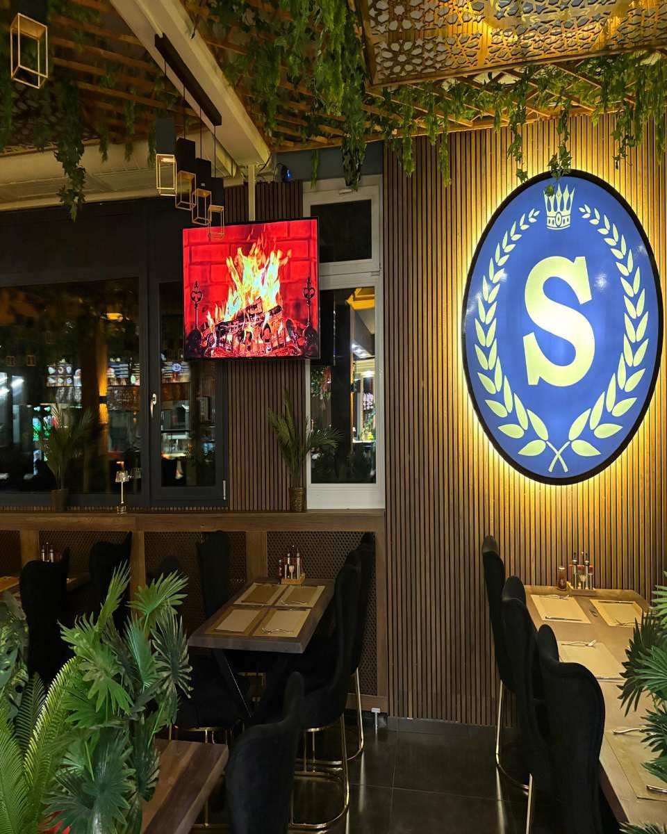 Restaurant-Ecke im Innenbereich mit blauem runden S-Schild, Holzdekor, Hängepflanzen und einem an der Wand montierten Fernseher, der ein Feuer zeigt