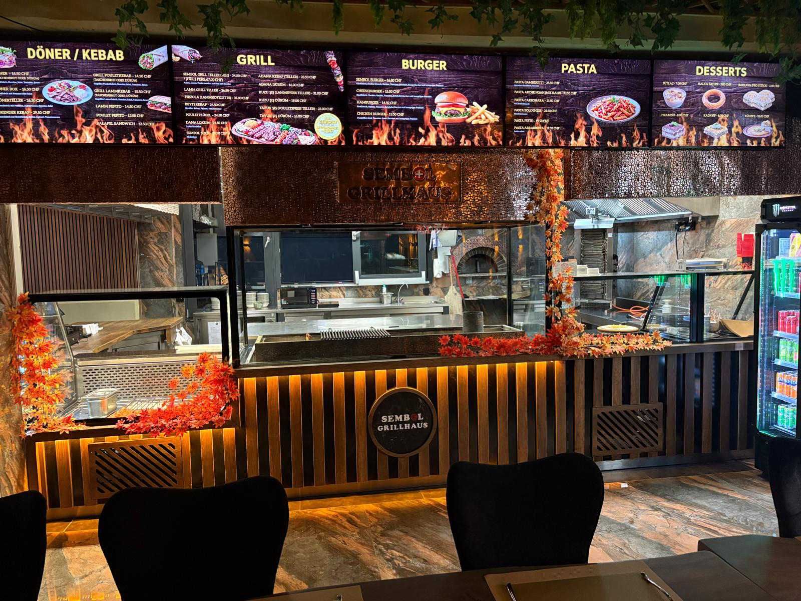Ein Imbissstand mit beleuchteten Menütafeln und einer gläsernen Auslage, gesehen von den Sitzplätzen im Freien.