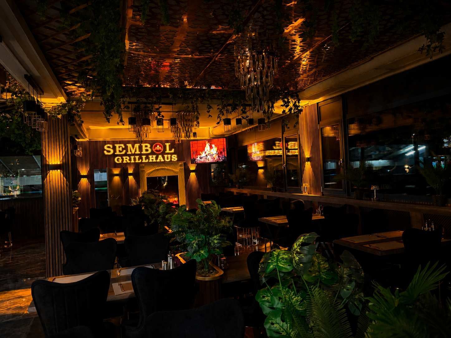 Die schwach beleuchtete Aussenterrasse des Restaurants mit Pflanzen, warmem, bernsteinfarbenem Licht und einem „SEMBOI“-Schild bei Nacht