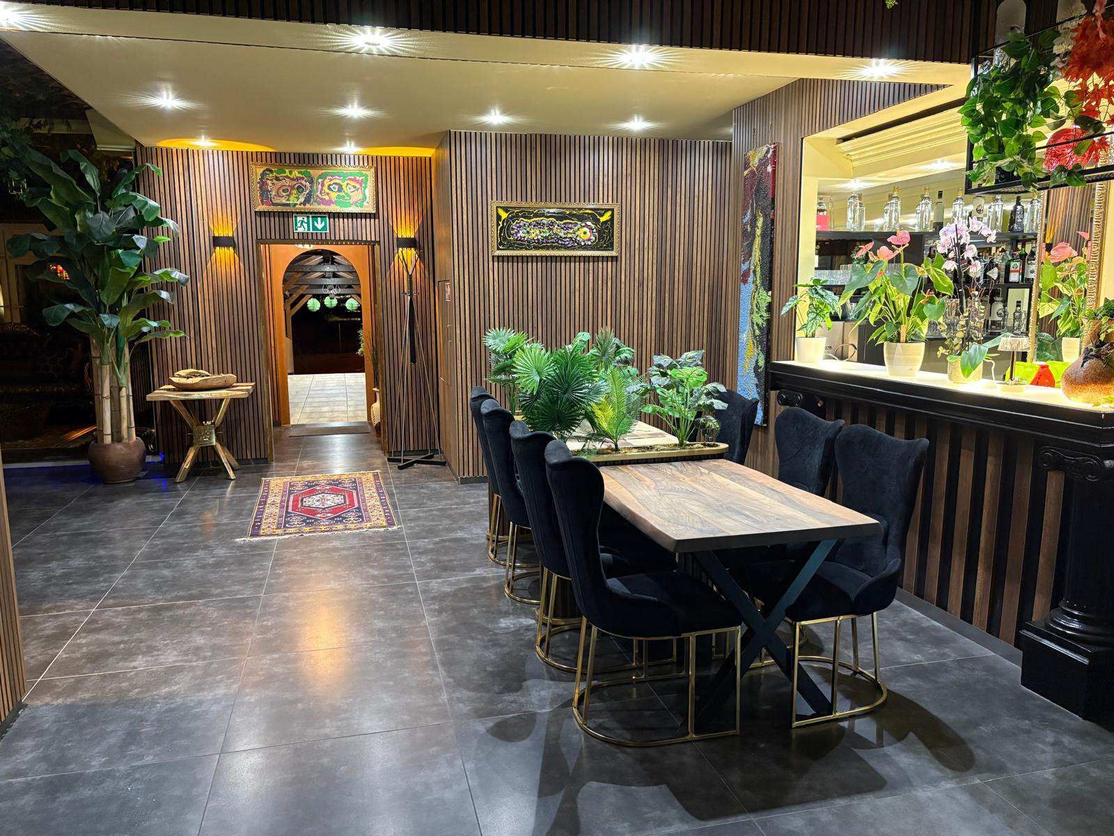 Gemütliches Restaurantinterieur mit dunklen Tischen, Stühlen, Holzelementen und üppigen grünen Pflanzen unter warmem Licht
