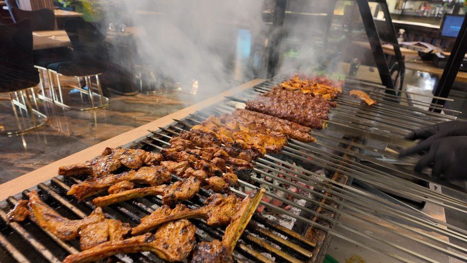 Gegrilltes Fleisch brutzelt über einem offenen Grill, Rauch steigt über den Spiessen auf.