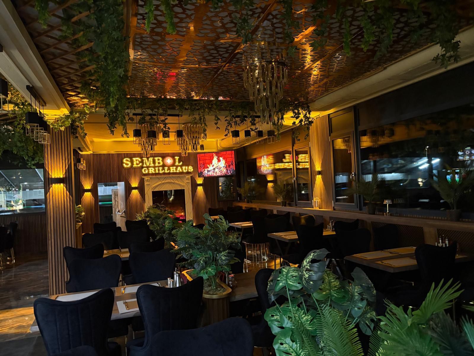 Gemütliche, pflanzenreiche Restaurantlounge mit dunklen Sitzgelegenheiten, bernsteinfarbener Beleuchtung und einer begrünten Decke.