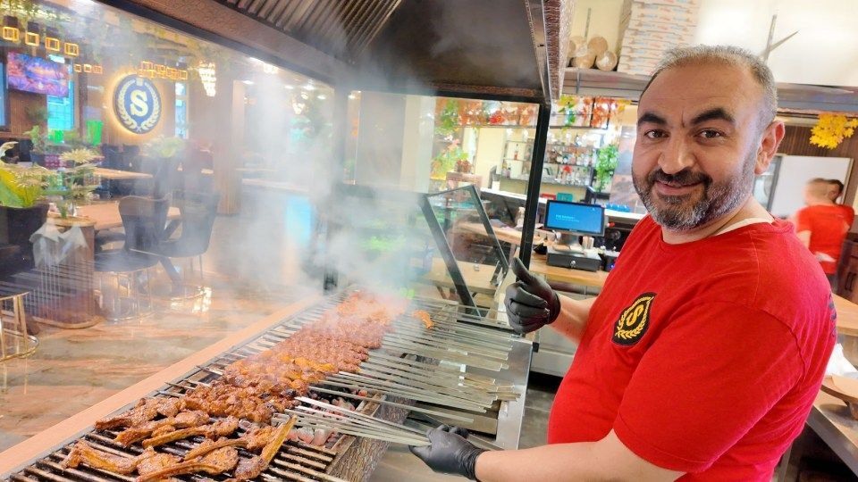 Ein Mann in einem roten Hemd grillt Spiesse an einem verrauchten Grillstand im Freien.