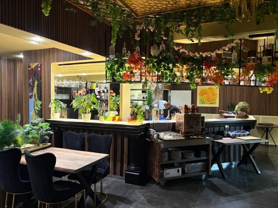 Stilvolles Barinterieur mit Hängepflanzen, verspiegelten Regalen, Holztischen und einer warm gelb beleuchteten Theke