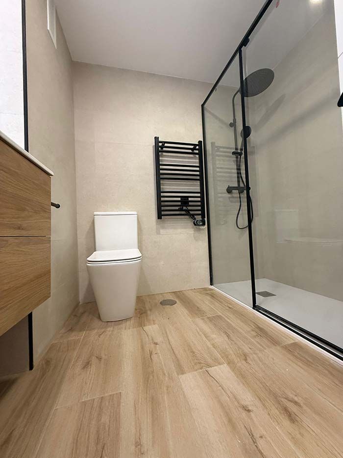 Baño moderno con ducha de cristal, inodoro, toallero y suelo de madera clara.