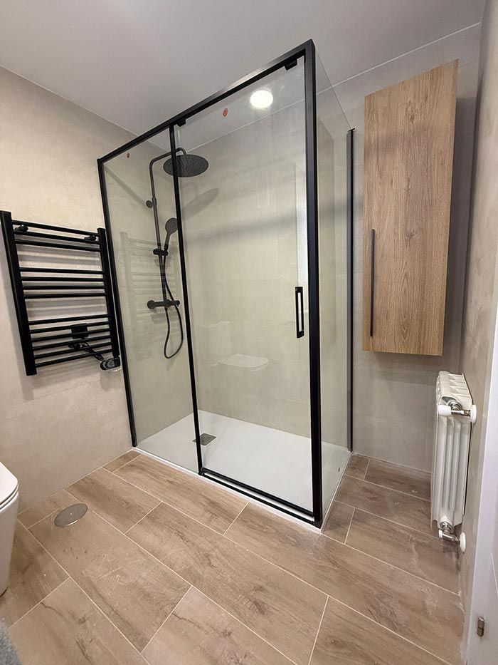 Baño moderno con ducha a ras de suelo de cristal, toallero negro, armario de madera y suelo de baldosas de madera clara.