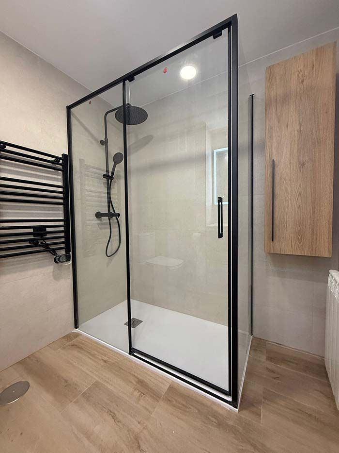 Baño moderno con ducha a ras de suelo de cristal, grifería negra, suelo de madera y armarios de pared.