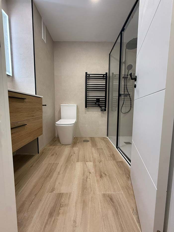 Baño moderno con lavabo de madera, inodoro, ducha de cristal y suelo con aspecto de madera.