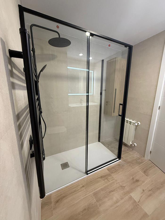 Ducha moderna a ras de suelo con marco negro y puertas de cristal, paredes de azulejos beige y suelo de baño con aspecto de madera.