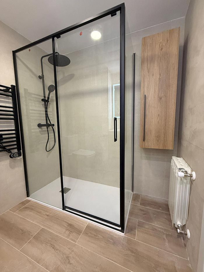 Baño moderno con cabina de ducha de cristal, paredes y suelo de azulejos beige, mueble de madera y radiador.