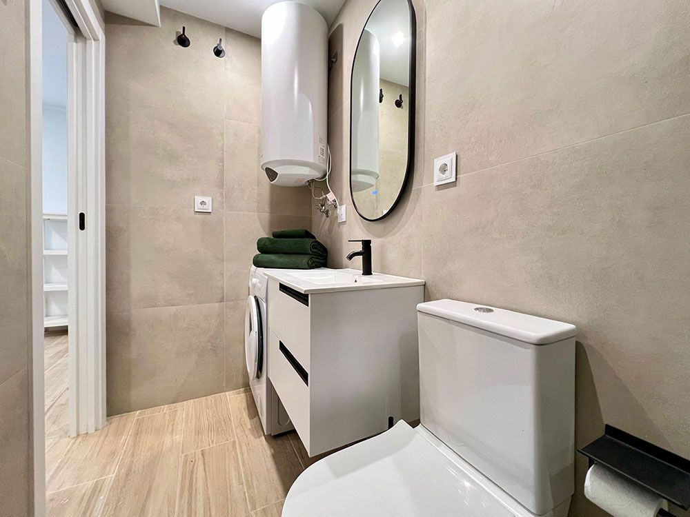 Baño pequeño de color beige con inodoro, lavabo, espejo y calentador de agua mural.