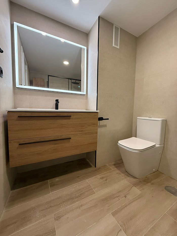 Baño moderno con tocador flotante de madera, espejo grande, lavabo e inodoro sobre suelo de madera clara.