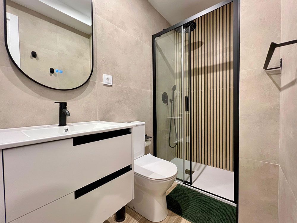Baño moderno con tocador blanco, espejo redondo, inodoro y ducha con mampara de cristal y detalles en negro.