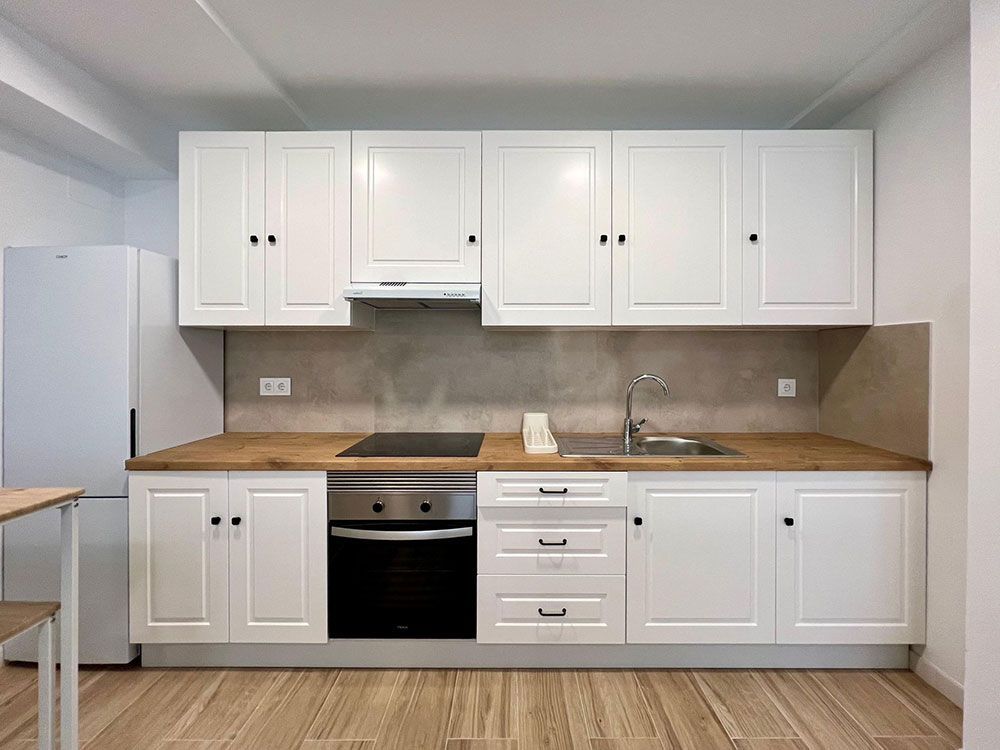Cocina blanca con armarios superiores e inferiores, encimera de madera, fregadero, cocina y refrigerador.