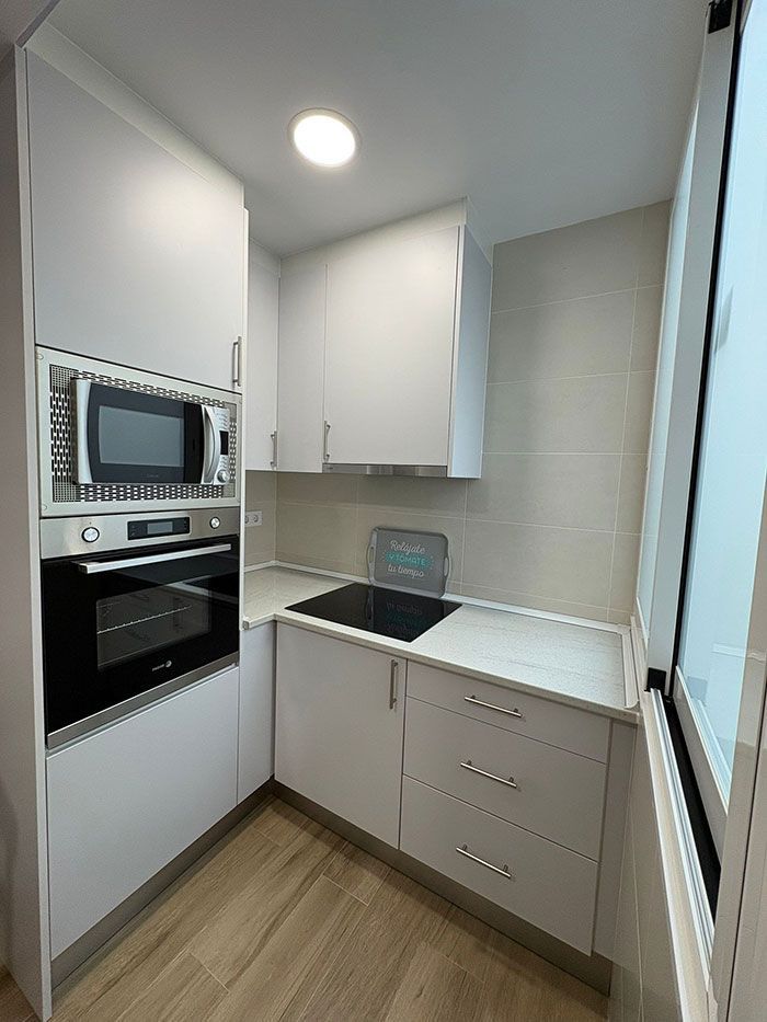Cocina compacta blanca con horno empotrado, microondas, placa de cocina negra y ventana junto a la encimera.