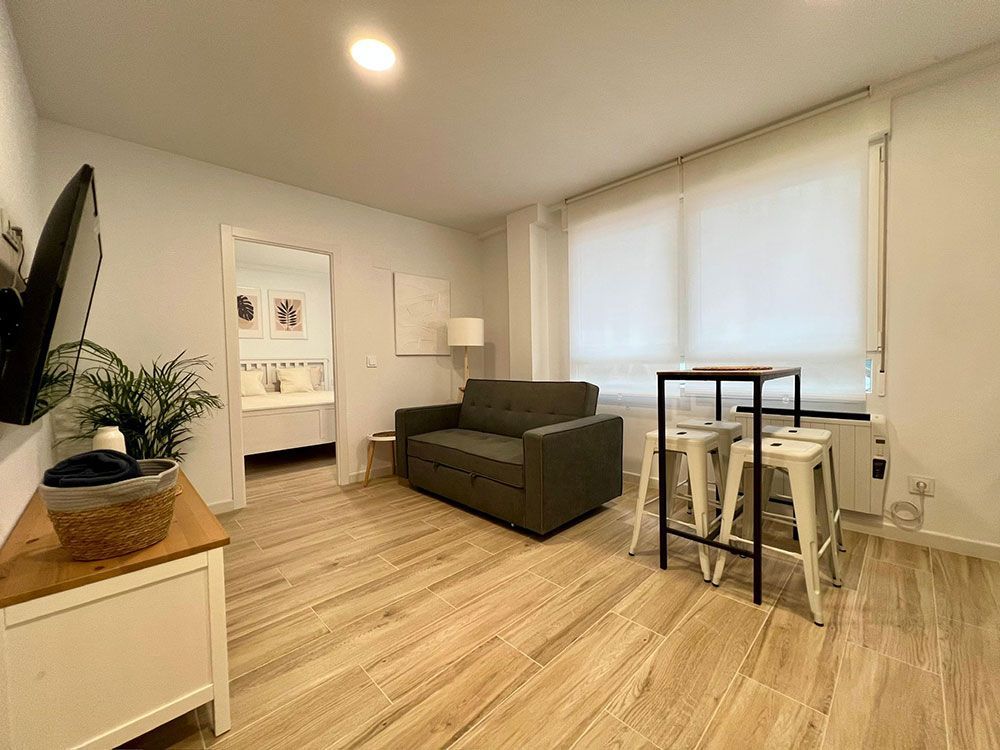 Sala de estar minimalista con sofá gris, mesa de comedor pequeña y persianas grandes sobre suelo de madera clara.
