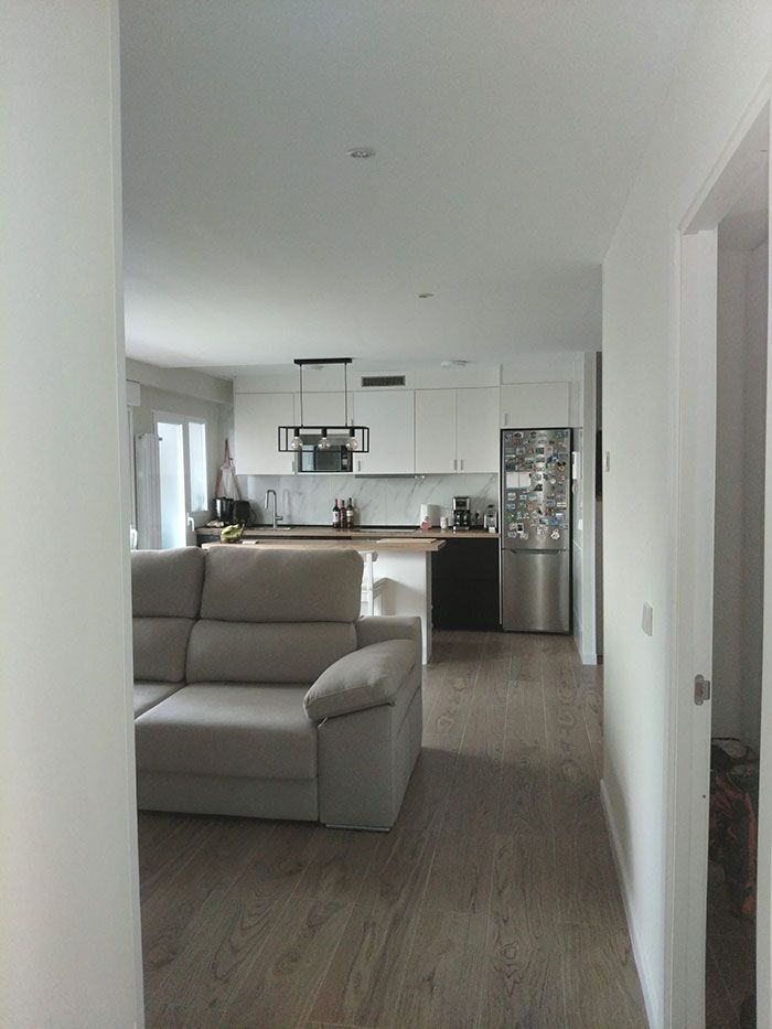 Salón moderno de planta abierta con sofá gris frente a una cocina blanca y refrigerador de acero inoxidable.
