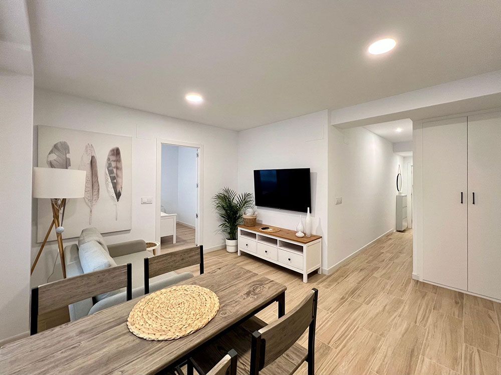 Luminoso salón-comedor moderno con paredes blancas, suelo de madera, televisión, sofá y mesa puesta.