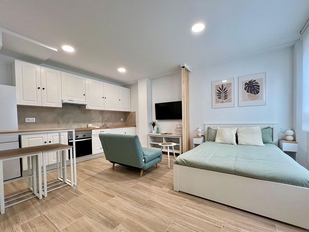 Apartamento tipo estudio luminoso con cocina blanca, silla color turquesa y cama junto a una zona de televisión.