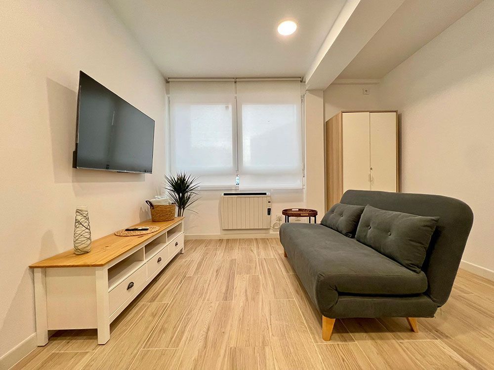 Sala de estar moderna con sofá gris, televisor montado en la pared, consola de madera y gran ventanal con persianas.