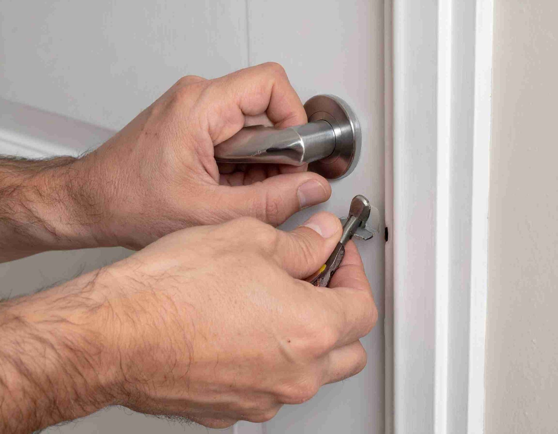 Manos que usan una llave para desbloquear una manija plateada en una puerta blanca en un entorno de pared beige.