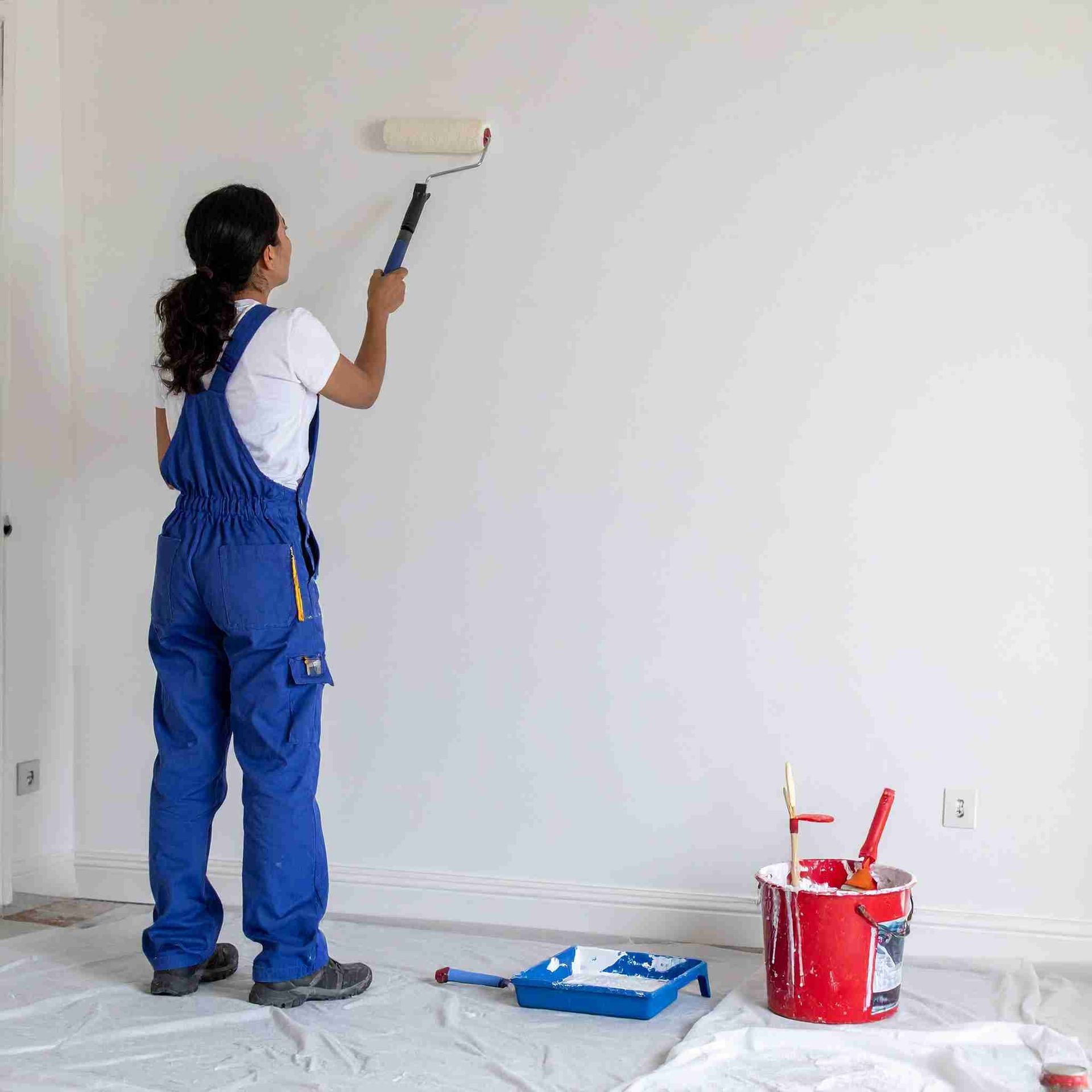 Mujer con mono azul pintando una pared blanca con un rodillo de pintura.