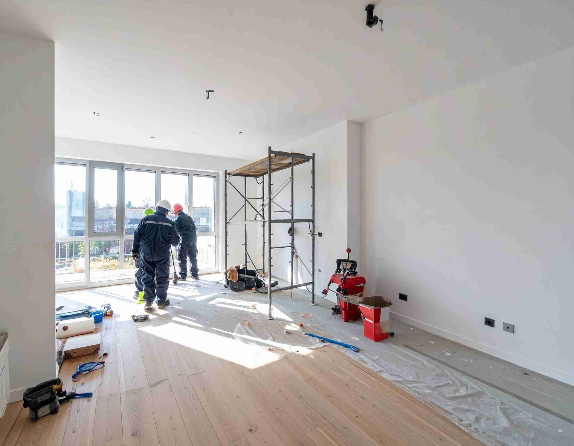 Obreros de la construcción dentro de un apartamento con andamios, herramientas y grandes ventanales; tonos neutros.