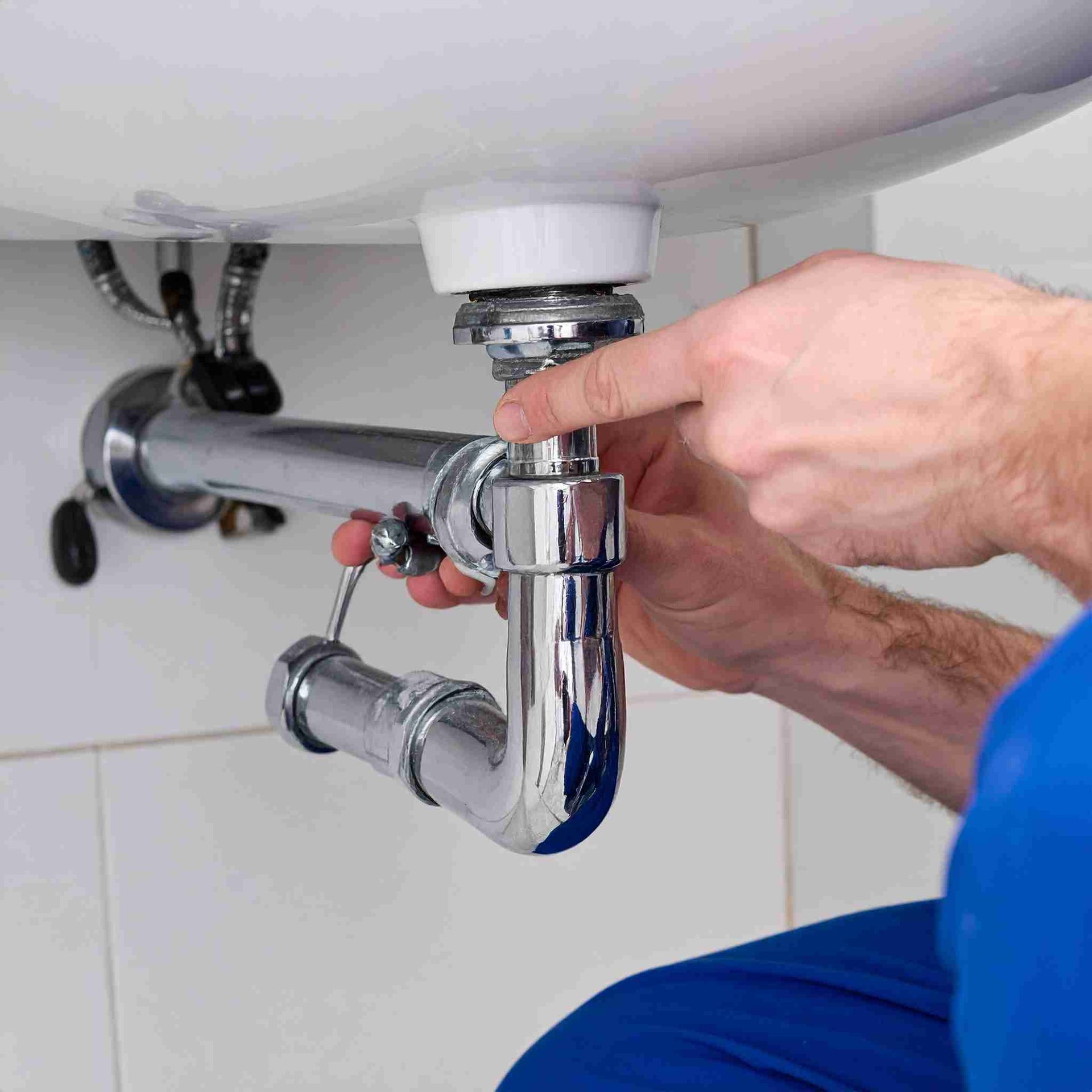 Un fontanero con camisa azul arregla una trampa P cromada debajo de un lavabo blanco en un baño.